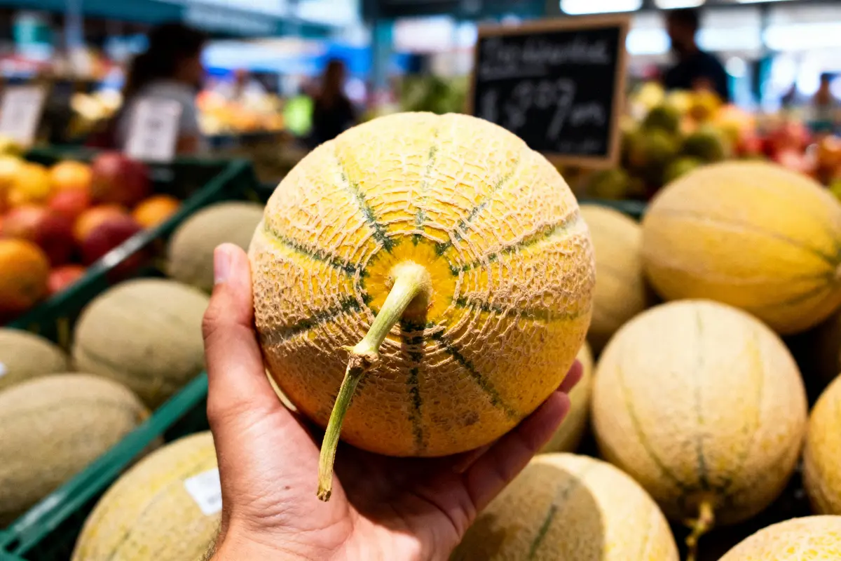 Main tenant un melon dans un magasin, étal de fruits en arrière-plan.