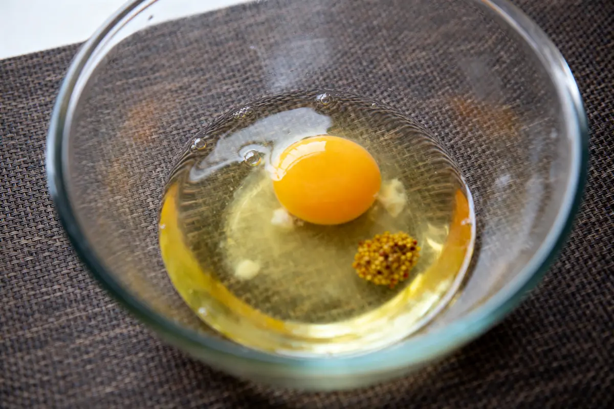 Bol transparent avec jaune d'œuf et moutarde à grains sur nappe textile marron.