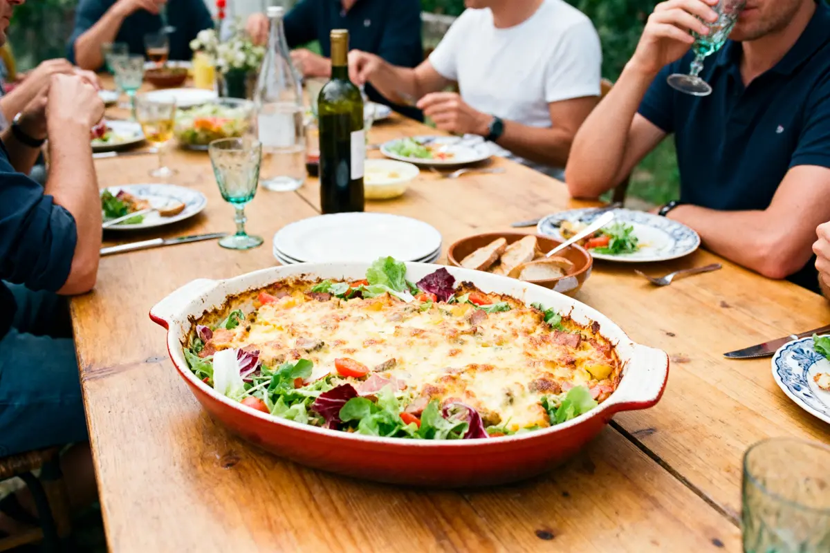 Un plat gratiné au fromage sur une table en bois, entouré de gens partageant un repas.