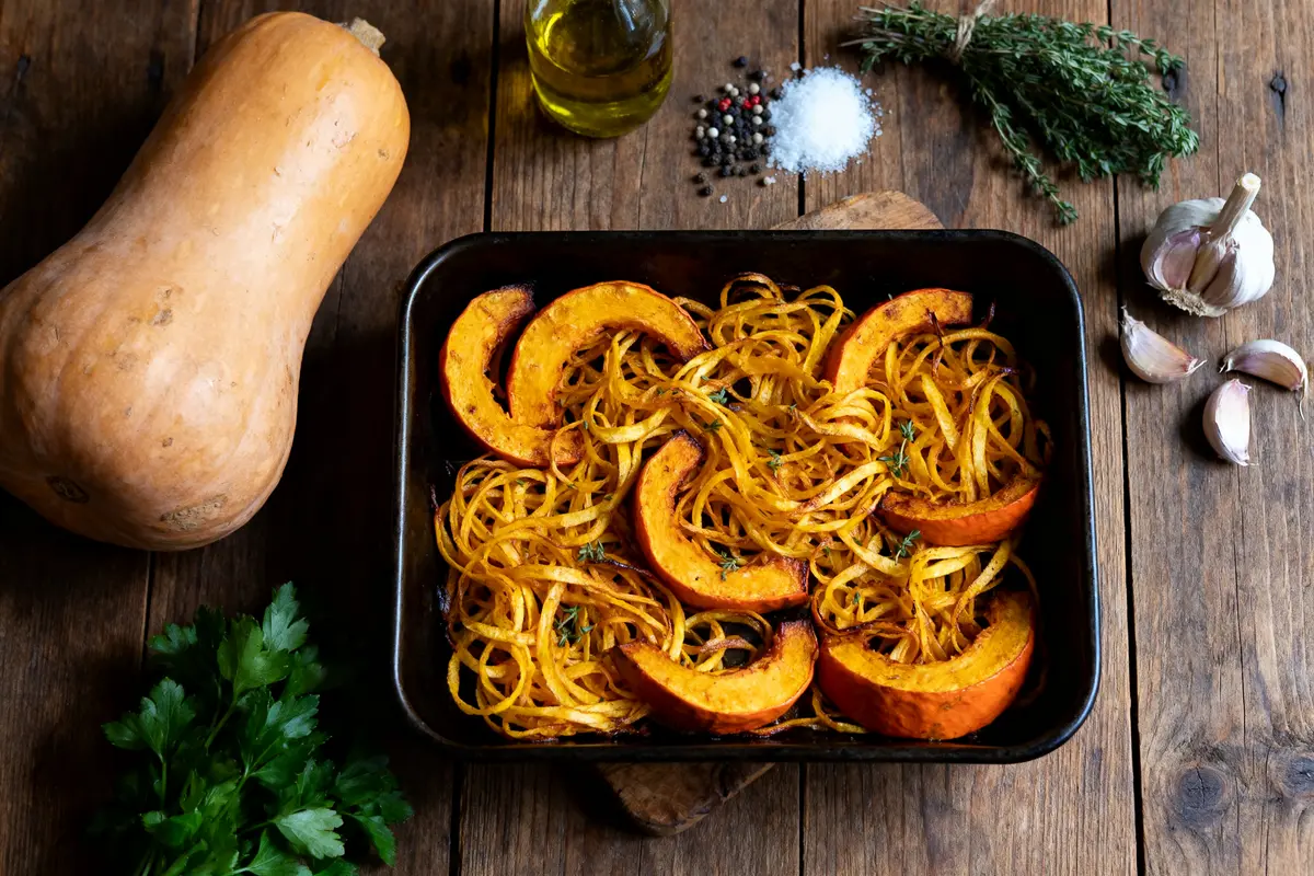 Tranches de courge et spirales rôties dans un plat, entourées d'ingrédients divers sur table en bois.