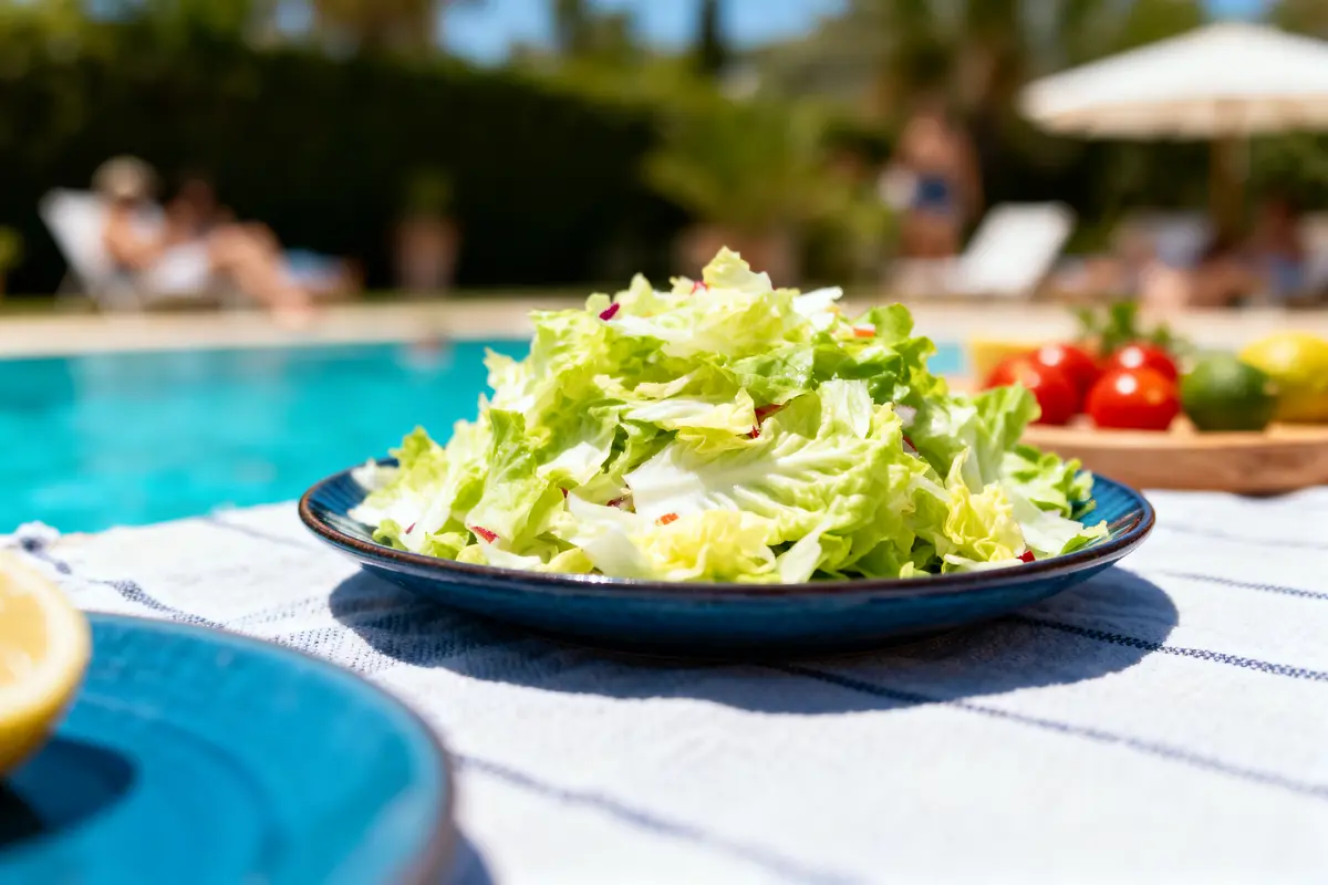 Assiette de salade verte sur table près d'une piscine, soleil, tomates, citron en arrière-plan flou.